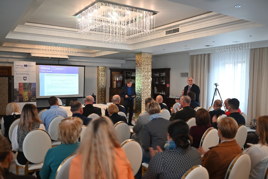 Grupa osób siedzi na krzesłach tyłem do zdjęcia i słucha prezentacji dwóch mężczyzn. Na ekranie wyświetla się slajd prezentacji . Obok ekranu znajduje się roll-up z logotypem Programu Skupina ľudí sedí na stoličkách chrbtom k kamere a počúva prezentáciu dvoch mužov. Na obrazovke je zobrazená prezentácia. Vedľa obrazovky je rozvinutý banner s logom programu.