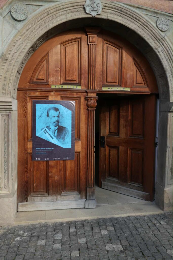 Drewniane dwuskrzydłowe drzwi wejściowe do budynku. Na nich zawieszony plakat ze zdjęciem Michała Mięsowicza i informacją o otwarciu wystawy dotyczącej jego działalności Drevené dvojité dvere vedúce do budovy. Na nich visí plagát s fotografiou Michała Mięsowicza a informáciami o otvorení výstavy jeho diel.