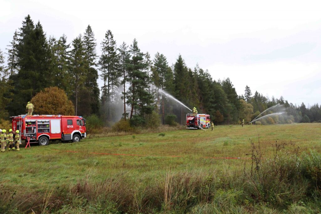 Dwa wozy strażackie stoją na skraju lasu. Z wozów leją się strumienie wody bezpośrednio na las. Dve hasičské autá stoja na okraji lesa. Z motorov sa priamo na les valia prúdy vody.