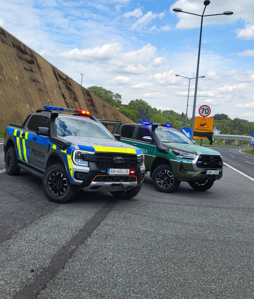 Dwa samochody policyjne typu pick-up zaparkowane na poboczu asfaltowej drogi. W tle znaki drogowe i duży spadek terenu Dve policajné dodávky zaparkované na okraji asfaltovej cesty. V pozadí dopravné značky a strmý svah.