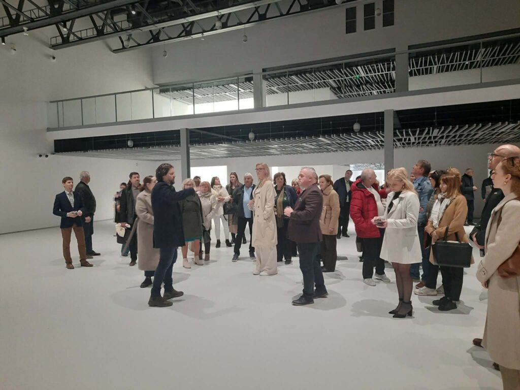 Grupa kilkudziesięciu mężczyzn i kobiet stoi w muzeum i słucha mężczyzny, który wskazuje ręką w prawą stronę Skupina niekoľkých desiatok mužov a žien stojí v múzeu a počúva muža, ktorý ukazuje rukou doprava