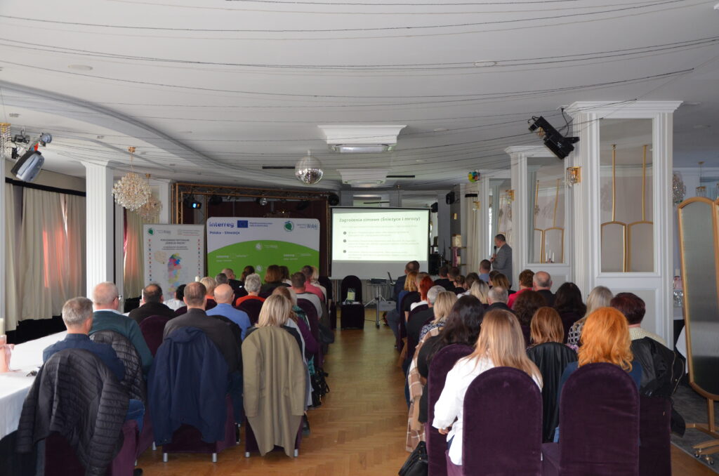 Kilkadziesiąt kobiet i mężczyzn siedzi na krzesłach na sali konferencyjnej i słucha prezentacji. Przed ekranem z prezentacją stoi mężczyzna z mikrofonem Niekoľko desiatok žien a mužov sedí na stoličkách v konferenčnej miestnosti a počúva prezentáciu
