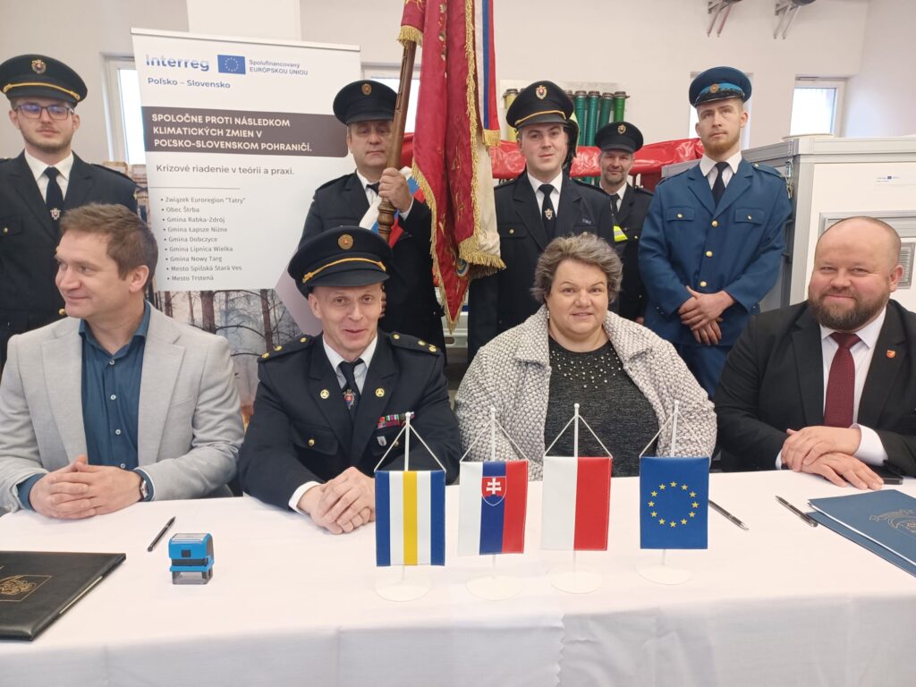 Trzech mężczyzn i jednak kobieta siedzą przy stole. Przed nimi ułożone flagi Unii Europejskiej, Polski i Słowacji. Za nimi stoi 5 strażaków w mundurach i ze sztandarem. Obok strażaków roll-up Pri stole sedia traja muži a jedna žena. Pred nimi sú rozložené vlajky Európskej únie, Poľska a Slovenska. Za nimi stojí päť hasičov v uniformách s vlajkou. Vedľa hasičov je roll-up