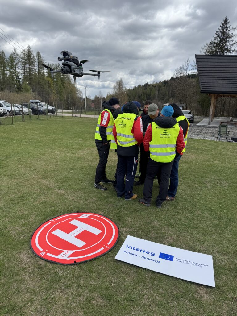 Kilka osób w kamizelkach stoi w kółku i rozmawia na polanie. Nad nimi lata dron. Przed nimi jest ustawione kółko z literą H jako cel lądowania drona i obok tablica z logotypem programu Niekoľko ľudí v vestách stojí v kruhu a rozpráva sa na lúke. Nad nimi lieta dron. Pred nimi je umiestnený kruh s písmenom H ako cieľ pristátia dronu a vedľa tabuľa s logom programu