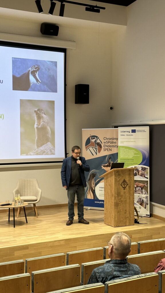Mężczyzna stoi na sali konferencyjnej, na podium z mikrofonem na tle ekranu Muž stojí v konferenčnej miestnosti na pódiu s mikrofónom pred obrazovkou