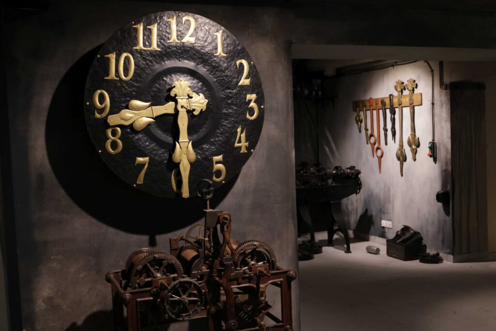 Wnętrze muzeum z wielkim zegarem pod którym znajduje się inny eksponat muzealny. W tle widać inne eksponaty Interiér múzea s veľkými hodinami, pod ktorými sa nachádza ďalší múzejný exponát. V pozadí sú vidieť ďalšie exponáty