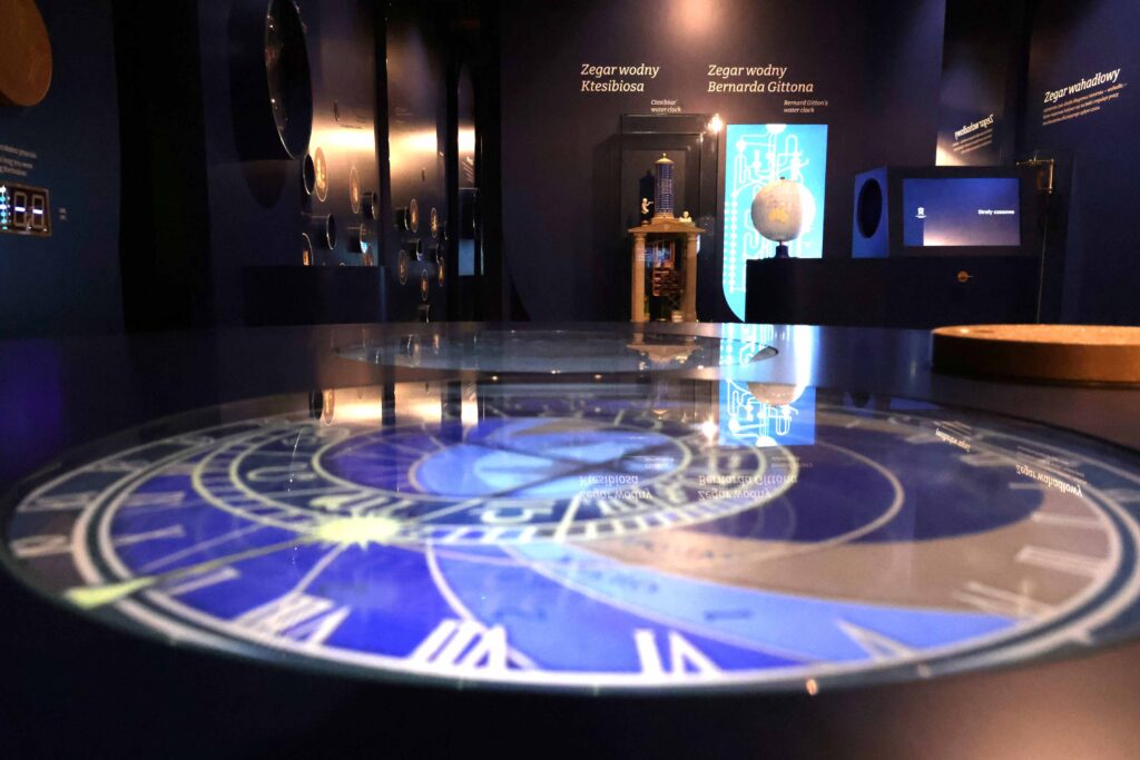 Przyciemnione wnętrze muzeum w którym widać różne interaktywne eksponaty związane z zegarami i czasem Zatienený interiér múzea, v ktorom sú vystavené rôzne interaktívne exponáty súvisiace s hodinami a časom