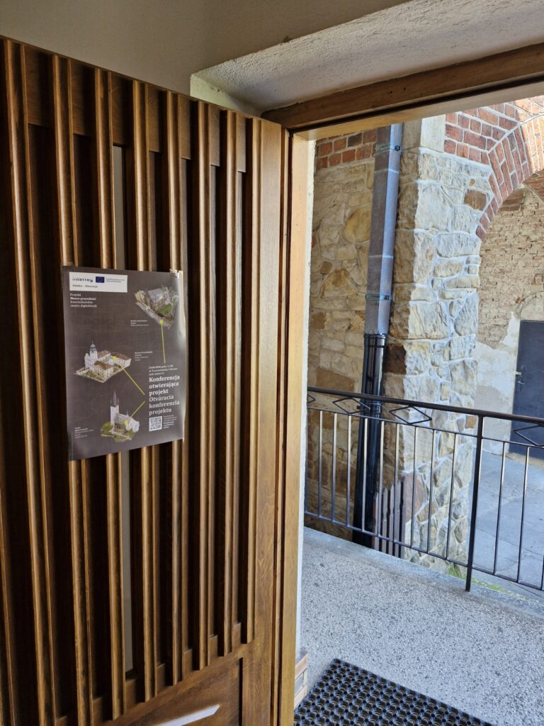 Plakat informacyjny na drewnianych drzwiach, zapowiadający konferencję otwierającą polsko-słowacki projekt Interreg „Wirtualna przeszłość franciszkańska – centra digitalizacji”. Na ciemnym tle plakatu widoczne są trójwymiarowe makiety trzech zabytkowych obiektów sakralnych połączone liniami oraz kod QR. W tle za otwartymi drzwiami fragment kamiennego muru z ceglanym łukiem i rynną. Informačný plagát na drevených dverách, ktorý pozýva na otváraciu konferenciu slovensko-poľského projektu Interreg „Virtuálna františkánska minulosť – digitalizačné centrá“. Na tmavom pozadí plagátu sú zobrazené trojrozmerné makety troch historických sakrálnych objektov prepojené líniami a QR kód. V pozadí za otvorenými dverami je vidieť časť kamenného múru s tehlovou klenbou a odkvapom.